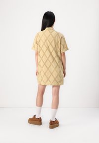 Donna con lunghi capelli neri in piedi di spalle, indossa un vestito corto a maniche corte beige traforato, calze bianche e scarpe con platform marroni su sfondo bianco.