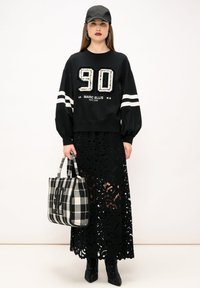 Felpa nera oversize con numero "90" in strass e righe bianche, indossata sopra una gonna in pizzo nera. Borsa a tracolla a quadri bianco e nero.