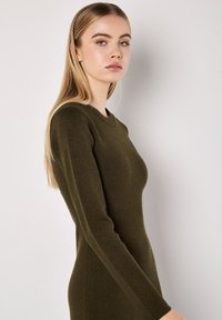 Olivegrünes langärmliges Kleid mit einer enganliegenden Silhouette, runder Ausschnitt und weicher Textur, seitlich betrachtet vor einem neutralen Hintergrund.