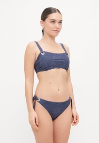 Sötétkék farmermintás bikini. A felsőrész széles pántokkal és ezüst kiegészítőkkel rendelkezik; a alsó rész állítható oldalsó kötések és sima felület jellemzi.