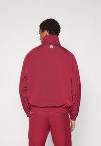 Casaco corta-vento bordô com gola alta, punhos elásticos e um logótipo nas costas. Feito de tecido leve e suave com um corte descontraído.