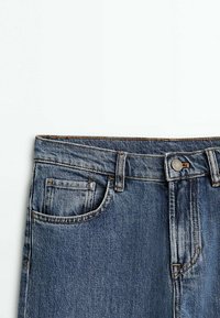 Blå denimjeans med midja, framficka, bältesömmar och metallknappslås på en vit bakgrund.