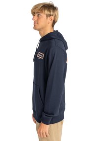 Sudadera azul marino con un bolsillo tipo canguro en la parte frontal, capucha ajustable con cordón y un detalle de parche en el pecho y la manga. Textura suave.