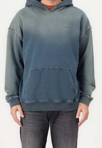 Hombre vestido con una sudadera azul-verde desteñida con bolsillo frontal y una camisa roja debajo, combinada con jeans gris oscuro, de pie contra un fondo blanco.