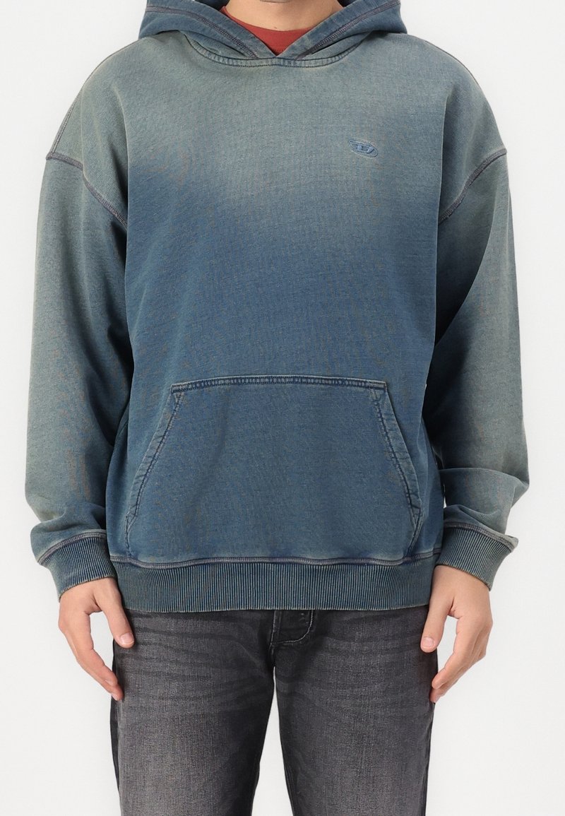 Hombre vestido con una sudadera azul-verde desteñida con bolsillo frontal y una camisa roja debajo, combinada con jeans gris oscuro, de pie contra un fondo blanco.