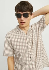 Chemise à manches courtes couleur ivoire avec un motif à carreaux rouges, dotée d'un col et d'une fermeture à boutons, portée avec des lunettes de soleil rectangulaires sombres.
