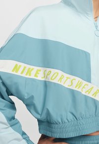 Persoană îmbrăcată într-o jachetă sport Nike Sportswear de culoare albastru deschis, scurtă, cu fermoar și talie elastică, cu mâna odihnită pe șold.
