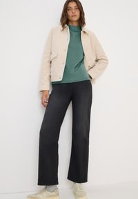Chaqueta texturizada beige con bolsillos delanteros, combinada con una camiseta verde oscuro y pantalones anchos negros, junto con zapatillas ligeras.
