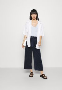 Vit överstor skjorta med knappar över en ljusblå linne, kombinerad med marinblå vida byxor och svarta sandals med spännen. Enkelt design.