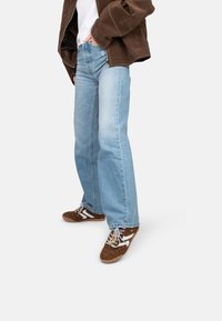 Jeans a gamba larga blu chiaro, t-shirt bianca, giacca scamosciata marrone, sneakers marroni con stampa leopardata e accenti bianchi, lacci grigi.