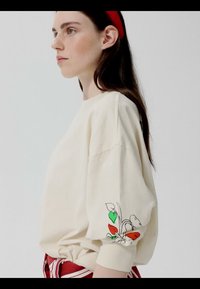 Giovane donna con felpa crema con motivo floreale rosso e verde sulla manica, indossa una fascia rossa per capelli e pantaloni a righe, guarda verso sinistra.