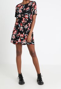 Schwarzes Blumenkleid mit roten und grünen Mustern, kurzen Ärmeln, elastischem Bund und ausgestelltem Rock. Kombiniert mit schwarzen Schnürstiefeletten.