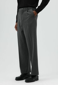 Grijze broek met een getailleerde pasvorm, voorzien van voorplooien, zijzakken en een knoopsluiting. Gecombineerd met zwarte leren schoenen.