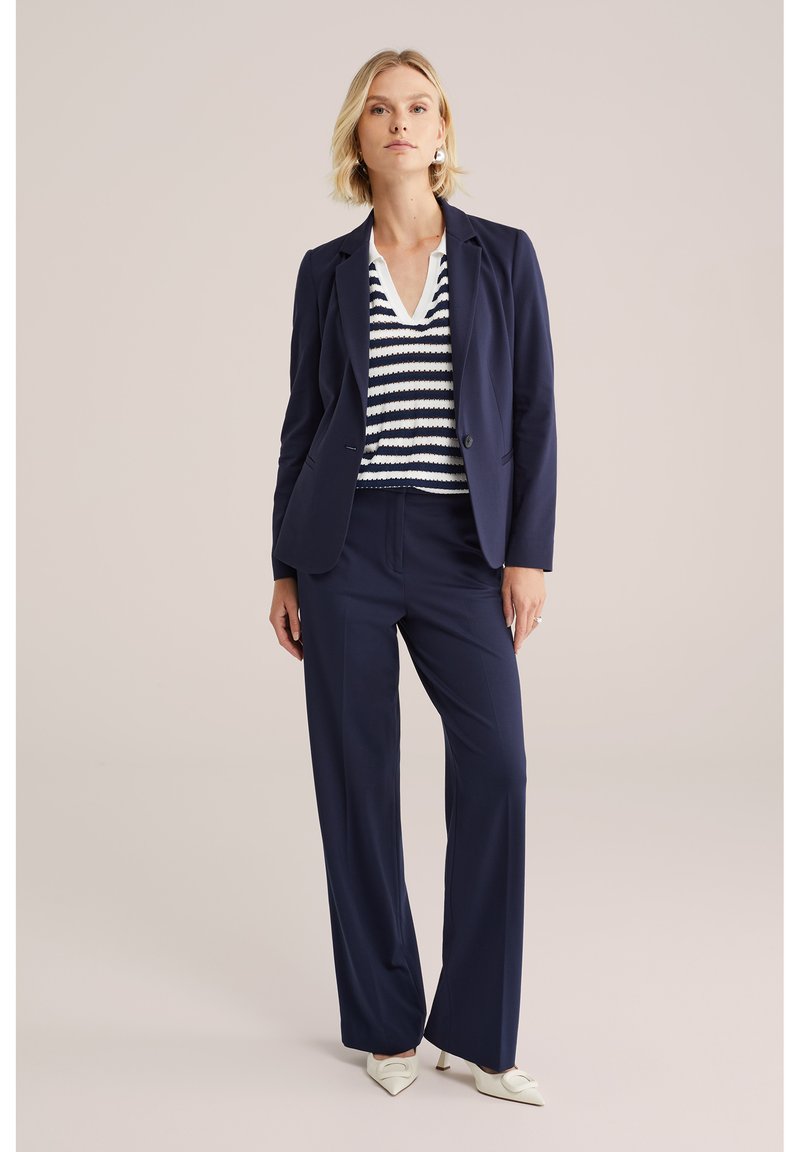 Femeie în picioare, purtând un sacou bleumarin și pantaloni, cu un tricou cu dungi albe și bleumarin în formă de V și pantofi cu toc ascuțit albi, pe un fundal simplu.