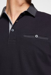 Černé polo tričko s kontrastním tečkovaným límcem, zapínáním na knoflíky a náprsní kapsou s dekorativním stehem. Měkká textura látky.