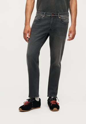 Homme portant un jean noir délavé, des baskets noires avec des lacets rouges et noirs, et des chaussettes gris clair, debout devant un fond uni.
