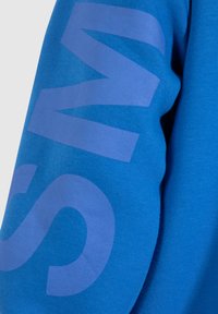 Sweat-shirt bleu avec une texture de maillot, arborant de grandes lettres violettes sur la manche. Le tissu semble doux et confortable.