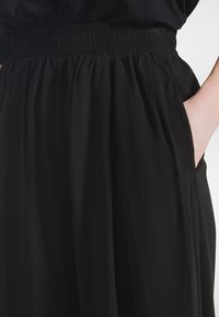 Mustat leveälahkeiset housut, joissa on joustava vyötärönauha ja sivutaskut. Housut ovat valmistettu sileästä, kevyestä kankaasta, jolla on virtaava tekstuuri.