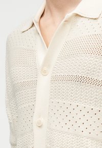 Cárdigan de punto en color crema con franjas de malla, que presenta un cuello y cierre de botones, mostrando un diseño texturizado y transpirable.