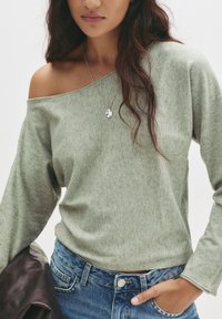Hellgrüner, schulterfreier Pullover mit langen Ärmeln und weicher Textur. Kombiniert mit hochtaillierten blauen Jeans aus Denim, die über eine Knopf- und Taschenverzierung verfügen.