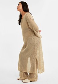 Langes beige Netzkleid mit weiten Ärmeln und seitlichen Schlitzen, kombiniert mit passenden weit geschnittenen Hosen und flachen Schuhen. Leichter und offen gewebter Stoff.