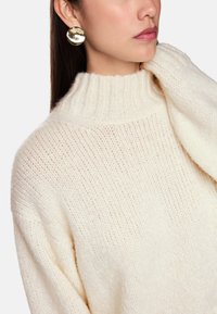 Cremefarbener Strickpullover mit hohem Rippkragen und strukturiertem Muster, kombiniert mit großen runden metallischen Ohrringen.