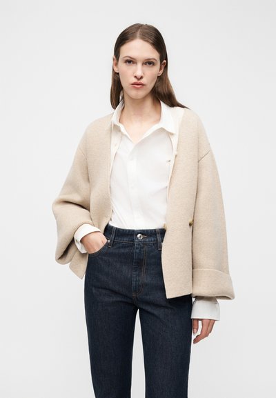 Γυναίκα που φοράει μπεζ oversized ζακέτα πάνω από λευκό πουκάμισο, σκούρο τζιν με ψηλή μέση, με το ένα χέρι στην τσέπη, στέκεται μπροστά σε απλό φόντο.