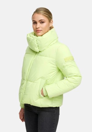 Vrouw in een lichtgroene gewatteerde jas met hoge kraag, handen in de zakken, en donkere jeans tegen een plain witte achtergrond.