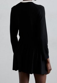 Person klädd i en svart långärmad klänning med veckad kjol, visad bakifrån mot en enfärgad ljusgrå bakgrund.