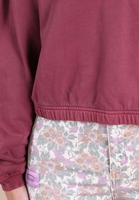 Sweatshirt bordeaux avec ourlet côtelé, associé à un pantalon à motif floral dans des teintes pastel de rose, gris et lavande. Comprend une étiquette de logo.