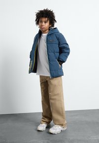 Giacca a vento blu con cappuccio, chiusura lampo frontale e polsini neri; abbinata a pantaloni beige e sneaker grigi. Design semplice con accenti a contrasto.