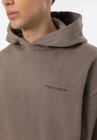 Rudas medvilnės džemperis su minkšta tekstūra ir laisvu siluetu. Priekyje yra ryškiai išsiuvinėtas „PEGAOOR“ logotipas.