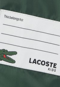 Zeleni tkaninski žig z napisom "To pripada:" v črni barvi, zelenim logotipom krokodila in napisom "LACOSTE KIDS" v črni barvi. Bela podlaga z obrobnim šivom.