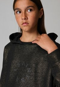 Sudadera negra con textura brillante, capucha y un logo redondo que dice "SAINT" en negro. El modelo muestra una mano en el cuello, destacando los detalles del diseño.