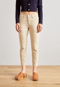 Jeans de mezcla de algodón beige con un corte ajustado, dos bolsillos frontales y cierre de botón, combinados con zuecos marrones sin hebilla con tacones de madera.