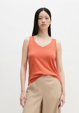 Jonge vrouw met lang zwart haar die een oranje mouwloos V-hals top en beige hoogtaillebroek draagt, staand tegen een effen witte achtergrond.