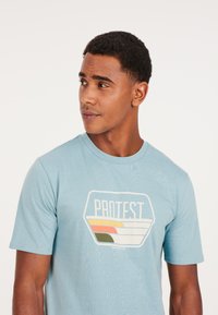 Camiseta de algodón azul claro con mangas cortas, con un diseño de logo geométrico en blanco, naranja, amarillo y verde en el frente.