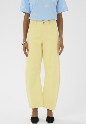 Osoba ubrana w żółte, szerokie spodnie z wysokim stanem, w połączeniu z jasnoniebieskim t-shirtem i czarnymi spiczastymi butami na jednolitym tle.