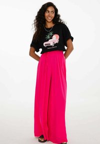 Žena s dlouhými kudrnatými vlasy, oblečená v černém tričku s grafikou a jasně růžových širokých kalhotách, stojí s rukama za zády.