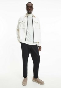 Giacca bianca con zip e due tasche frontali sopra un dolcevita beige chiaro, abbinata a pantaloni neri e scarpe beige con lacci.
