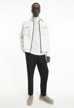 Giacca bianca con zip e due tasche frontali sopra un dolcevita beige chiaro, abbinata a pantaloni neri e scarpe beige con lacci.