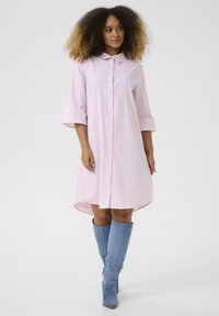 Abito camicia a righe rosa con colletto, chiusura frontale a bottoni e maniche arrotolate. Indossato con stivali blu fino al ginocchio.