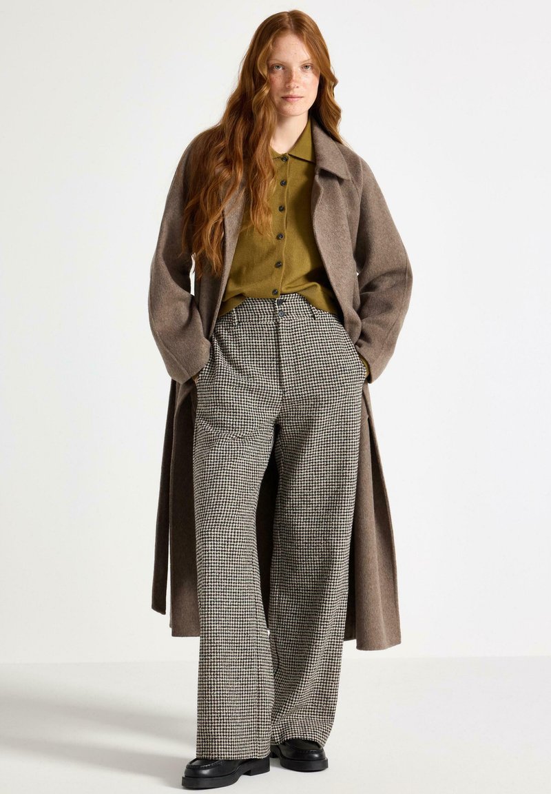 Giovane donna con lunghi capelli rossi, vestita con un lungo cappotto marrone, una camicia verde oliva con bottoni, pantaloni a quadri a gamba larga e scarpe nere, in piedi con le mani nelle tasche.