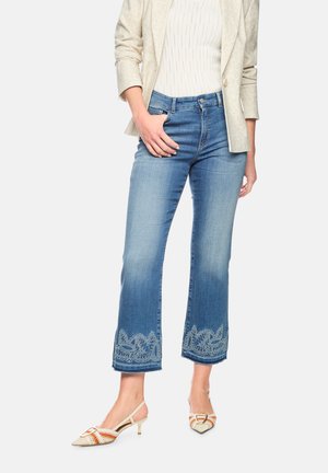 Frau trägt verblasste, blaue Cropped-Jeans mit bestickten Säumen, beige Blazer, weißes Oberteil und spitze Slingback-Pumps mit kleinen Kitten-Heels.