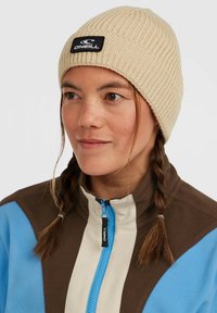 Bež õmblusega beanie, millel on sooniline tekstuur ja must logo silt. Kanda pruunide, siniste ja kreemjate toonide särgil, millel on tõmblukk ja seisev krae.