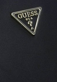 Materiale nero con texture e logo triangolare metallico di Guess USA che presenta un punto interrogativo e una freccia all'interno del triangolo.