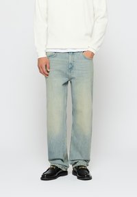 Világoskék farmernadrág laza szabással, látható fakulással, klasszikus öt zsebes dizájnnal, fehér hosszú ujjú felsővel és fekete loaferekkel párosítva.