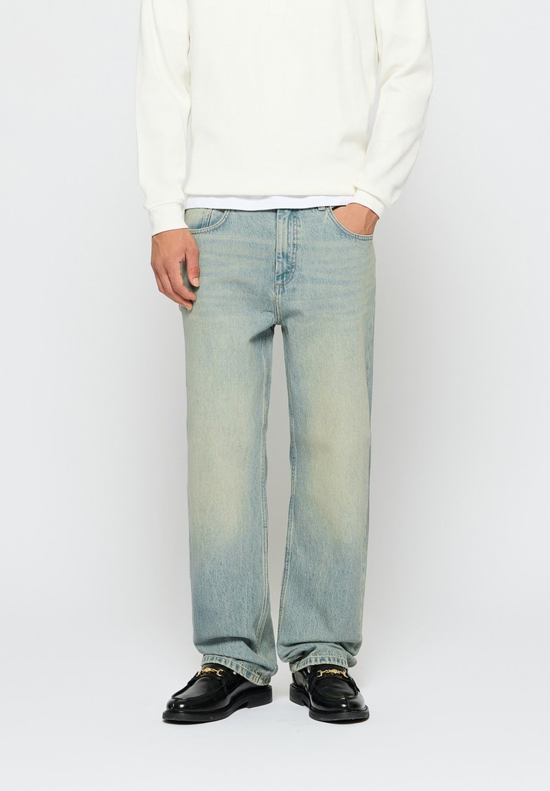 Világoskék farmernadrág laza szabással, látható fakulással, klasszikus öt zsebes dizájnnal, fehér hosszú ujjú felsővel és fekete loaferekkel párosítva.