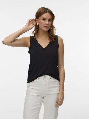 Femme aux cheveux châtain clair portant un haut noir sans manches à col en V et un pantalon blanc, touchant son oreille gauche avec la main droite.