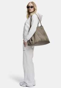 Lys grå hettegenser med hette, matchende bukser og en stor brun totebag. Teksturert stoff med synlige sømmer og sting.